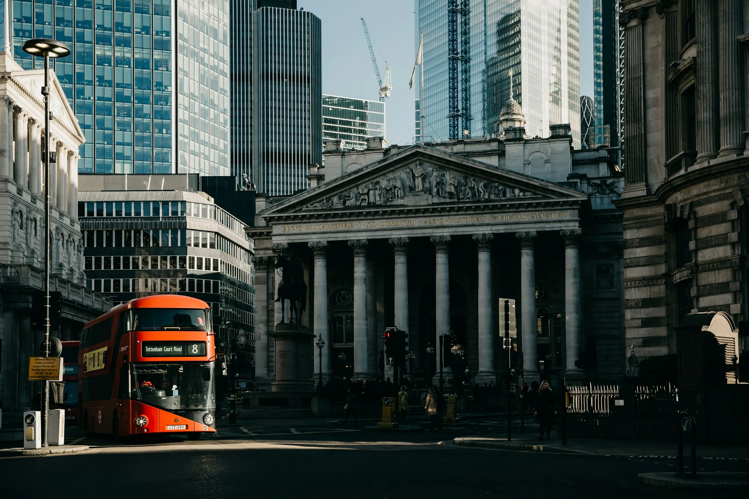 City of London street scene with the Royal Exchange columned façade, a red London bus passing in front.